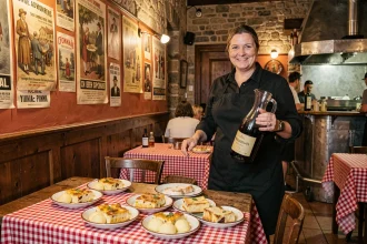 Serveuse dans un bouchon lyonnais traditionnel portant un carafe de Beaujolais avec des plats typiques sur table à carreaux rouge et blanc
